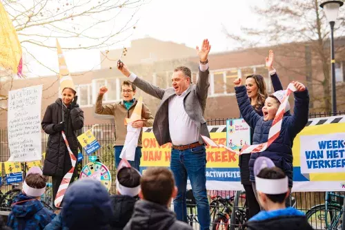 Een groep volwassen en kinderen staat op een schoolplein bij de feestelijke opening van de Week van het Verkeer. Wethouder Coen Lageveen staat in het midden en steekt beide armen in de lucht terwijl een lint omhoog wordt gehouden. Voor het podium staan kinderen met papieren hoofdbandjes. Op de achtergrond hangen kleurrijke banners over de Week van het Verkeer en staat een rad met verkeersvragen.
