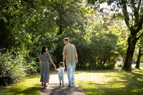 Een gezin dat wandelt in een park. Je ziet de moeder en vader, en daartussen in hun kind van grofweg een jaar of 2. Ze lopen in een groen park in Purmerend. Ze lopen weg van de kijker 