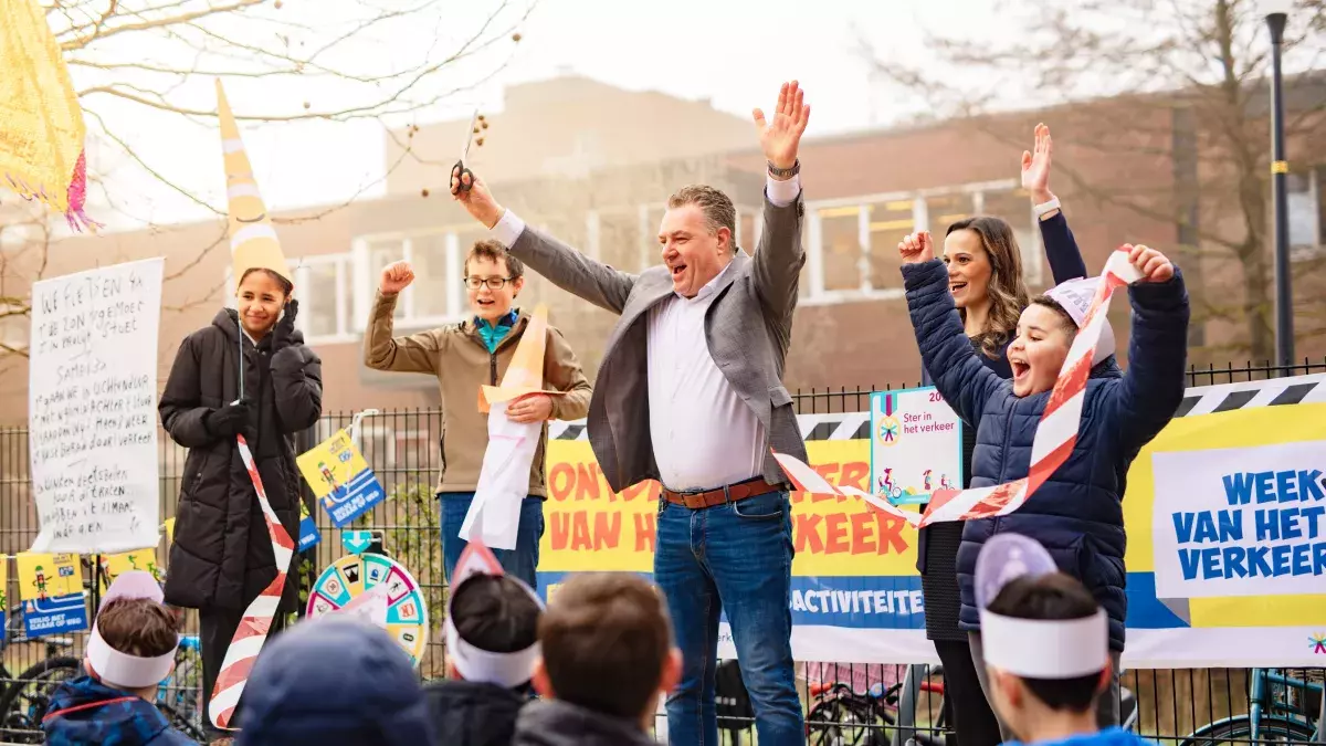 Een groep volwassen en kinderen staat op een schoolplein bij de feestelijke opening van de Week van het Verkeer. Wethouder Coen Lageveen staat in het midden en steekt beide armen in de lucht terwijl een lint omhoog wordt gehouden. Voor het podium staan kinderen met papieren hoofdbandjes. Op de achtergrond hangen kleurrijke banners over de Week van het Verkeer en staat een rad met verkeersvragen.