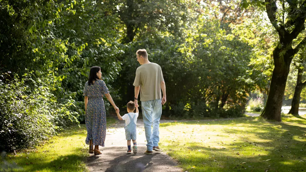 Een gezin dat wandelt in een park. Je ziet de moeder en vader, en daartussen in hun kind van grofweg een jaar of 2. Ze lopen in een groen park in Purmerend. Ze lopen weg van de kijker 